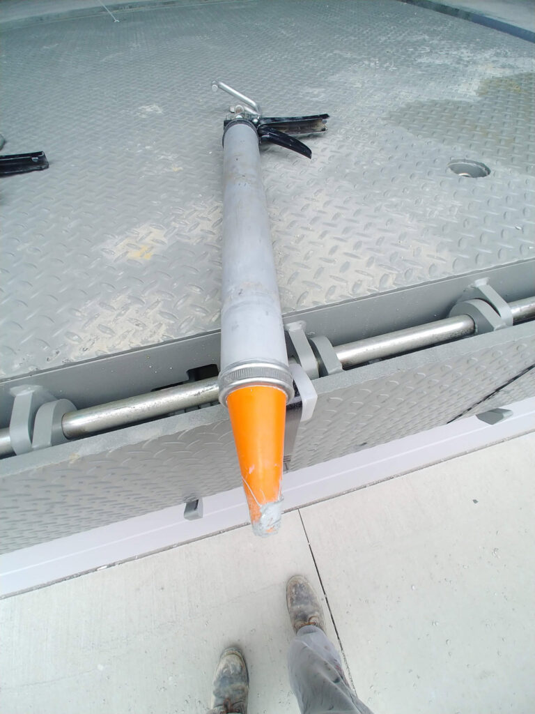 Caulking gun resting on a metal access hatch beside a concrete surface"