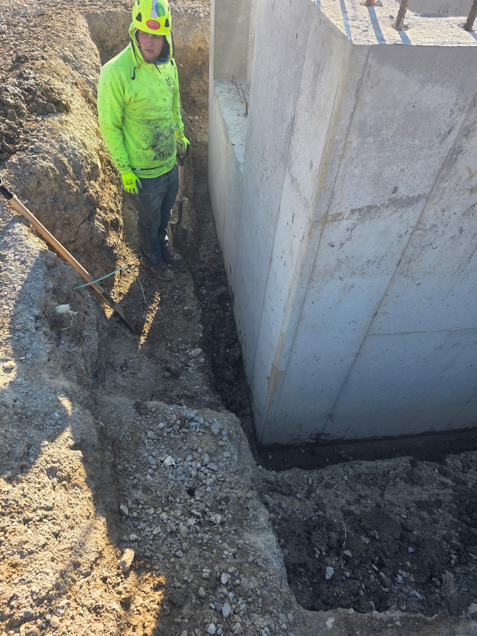 Worker beside a commercial concrete foundation wall in an open below-grade excavation before backfill.