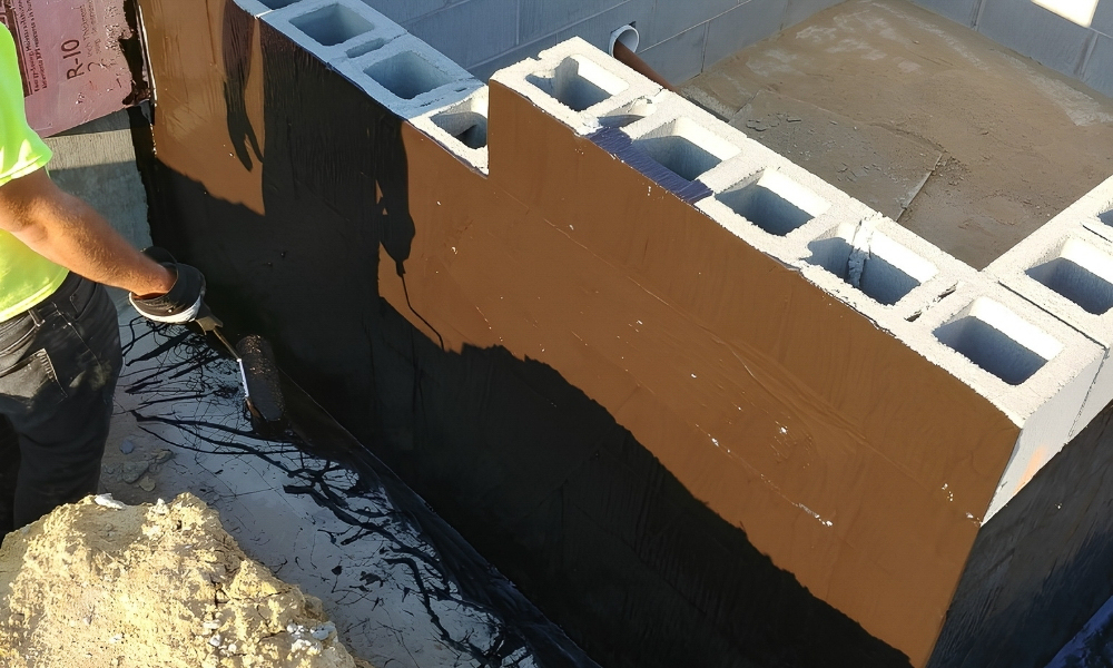 Worker applying a black coating to a commercial masonry foundation wall.