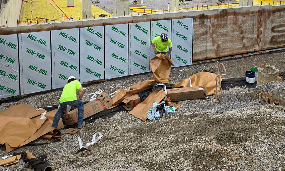 Workers installing sheet waterproofing membrane on a commercial concrete foundation wall.