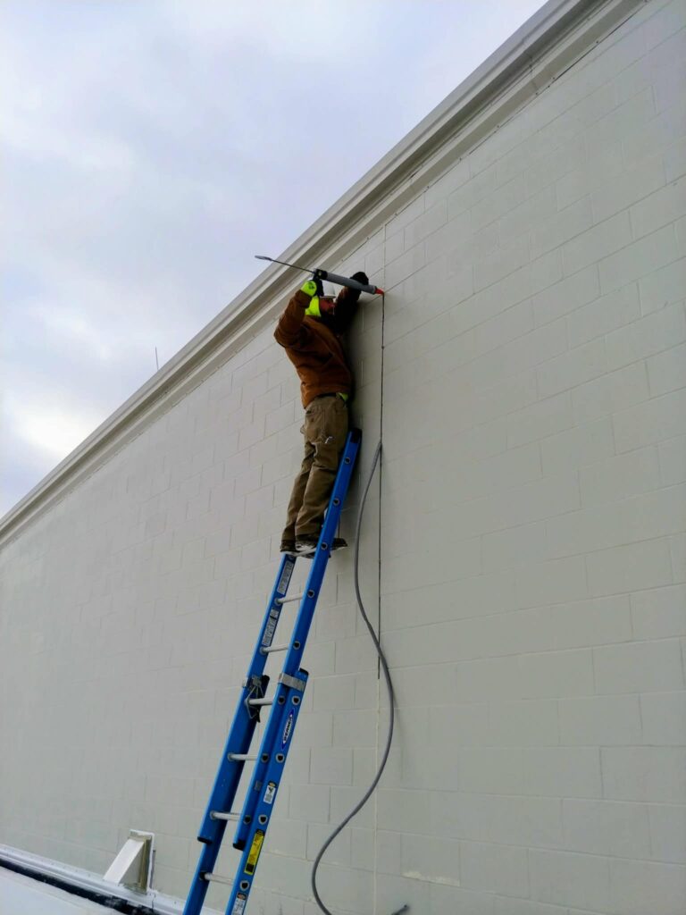 Worker on a blue ladder applying sealant along a vertical exterior masonry joint."