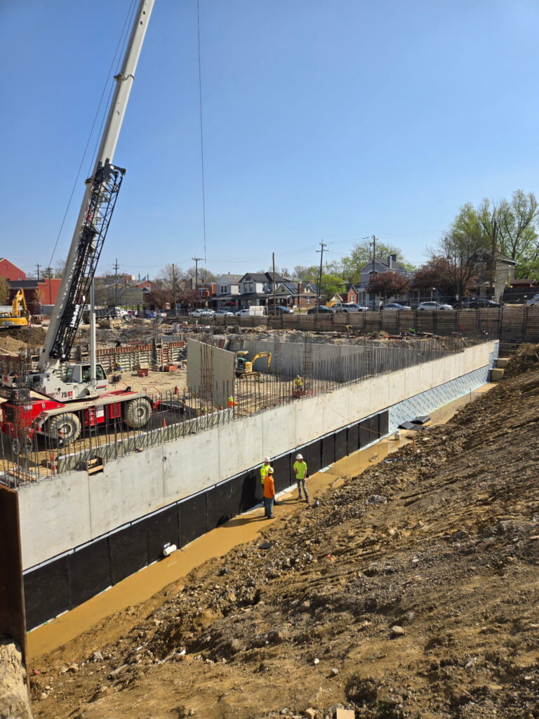 Crew beside a waterproofed concrete foundation wall before backfilling, with a crane in the background."